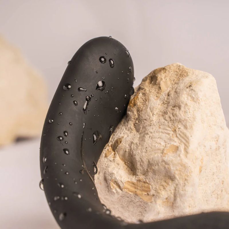 Black silicone cock and ball vibrator with droplets next to a textured rock surface.