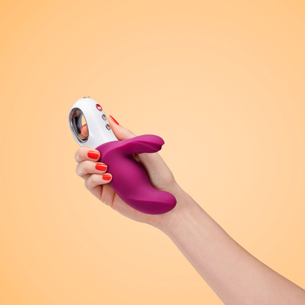 Hand holding Fun Factory MISS BI grape - Grape 17 cm USB Rechargeable Rabbit Vibrator against a peach background.