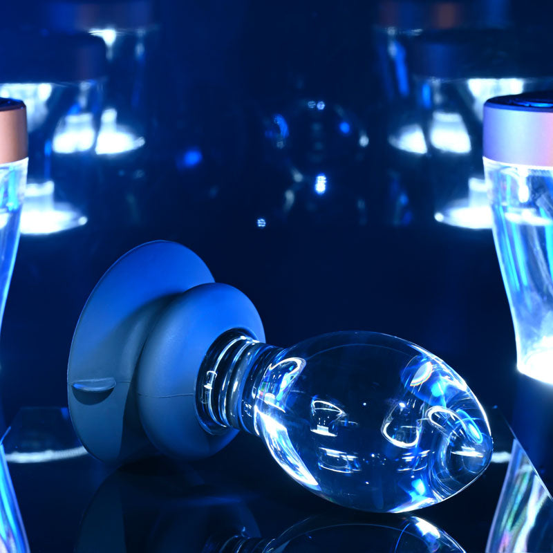 Gender X Crystal Ball -  Glass 13.6 cm Butt Plug with Suction Base on a reflective surface with blue lighting.