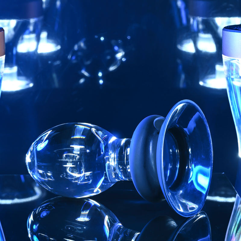 Gender X Crystal Ball - Glass 13.6 cm Butt Plug with Suction Base on a reflective surface with blue lights.