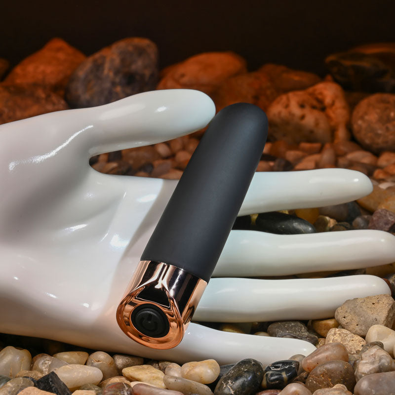 Gender X THE GOLD STANDARD - /Rose Gold 10 cm USB Rechargeable Bullet resting on a hand among decorative stones.