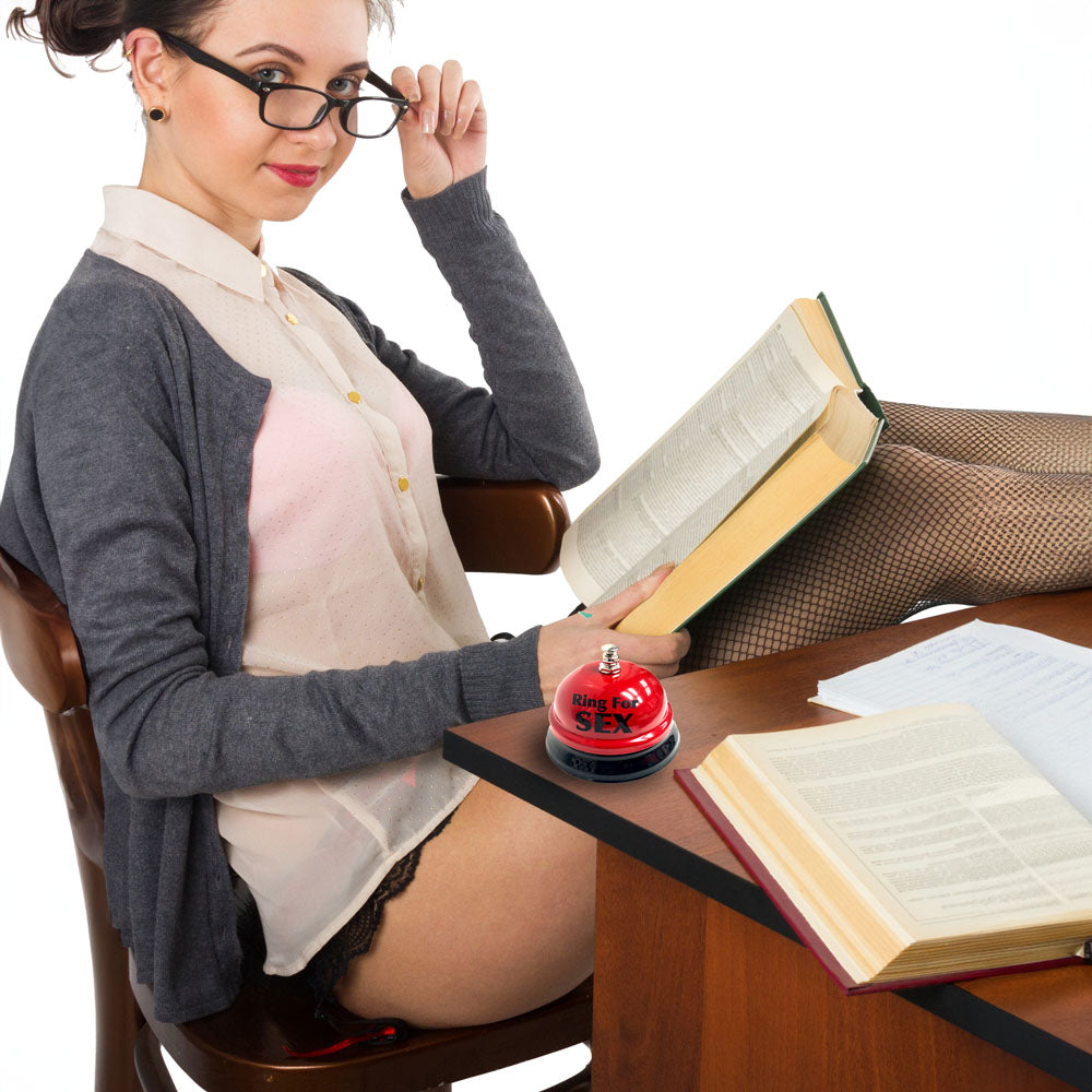 A woman in glasses with a Ring For Sex Table Bell - Novelty Bell on her desk, reading books in a playful pose.