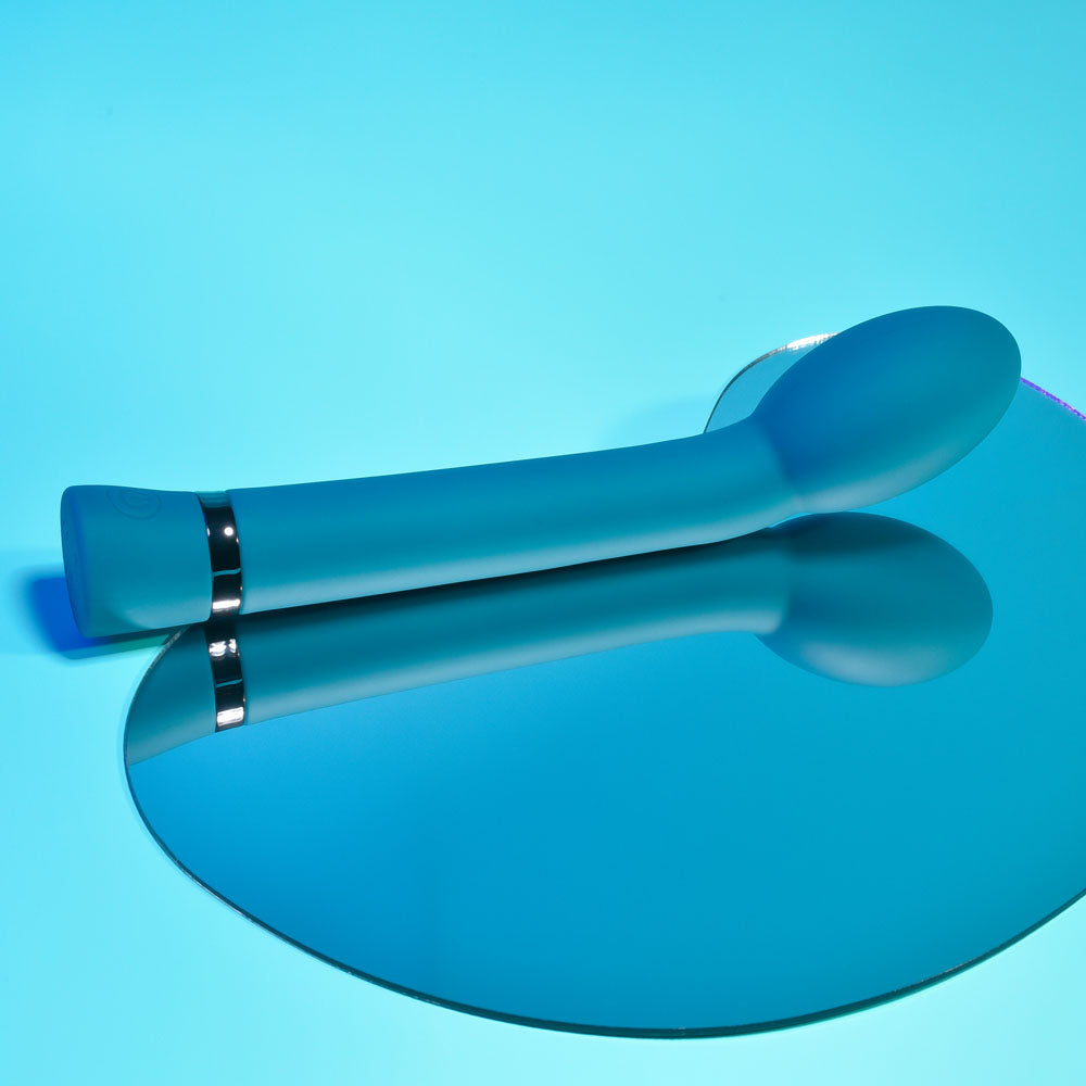 Playboy Pleasure ON THE SPOT G-Spot vibrator on a reflective surface against a blue background.