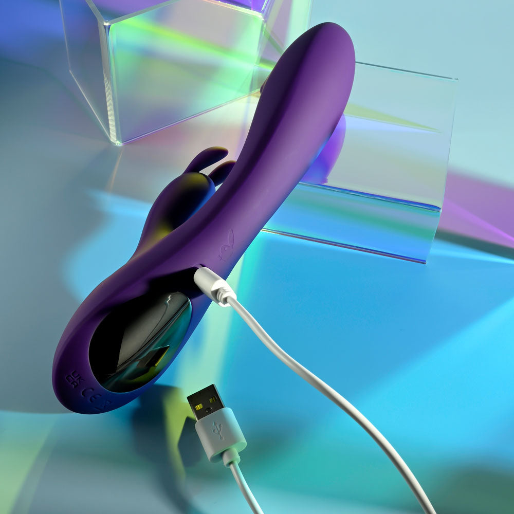 Playboy Pleasure ON REPEAT - 19.7 cm USB Rechargeable Rabbit Vibrator charging against a colorful background.