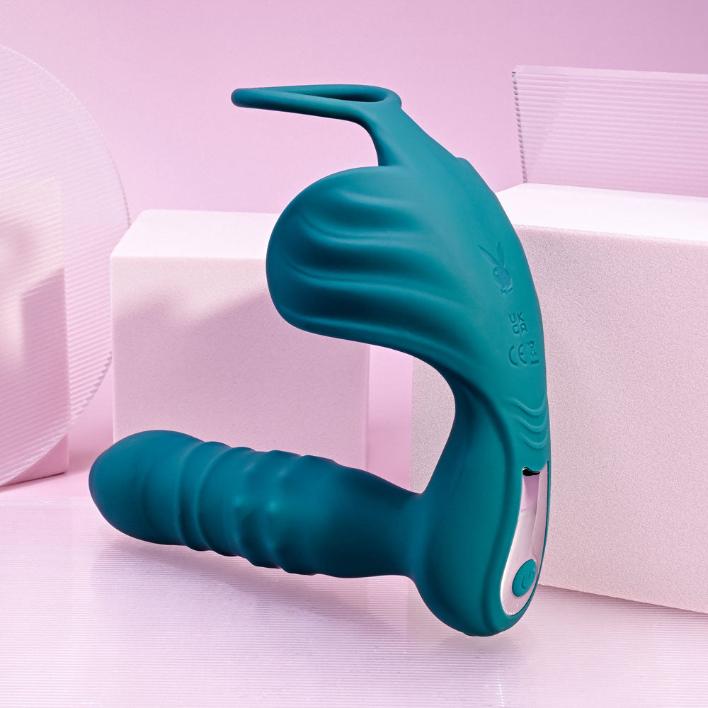 Playboy Pleasure BRING IT ON - Teal USB Rechargeable Thrusting Anal Plug with Cock Ring on a pink background.