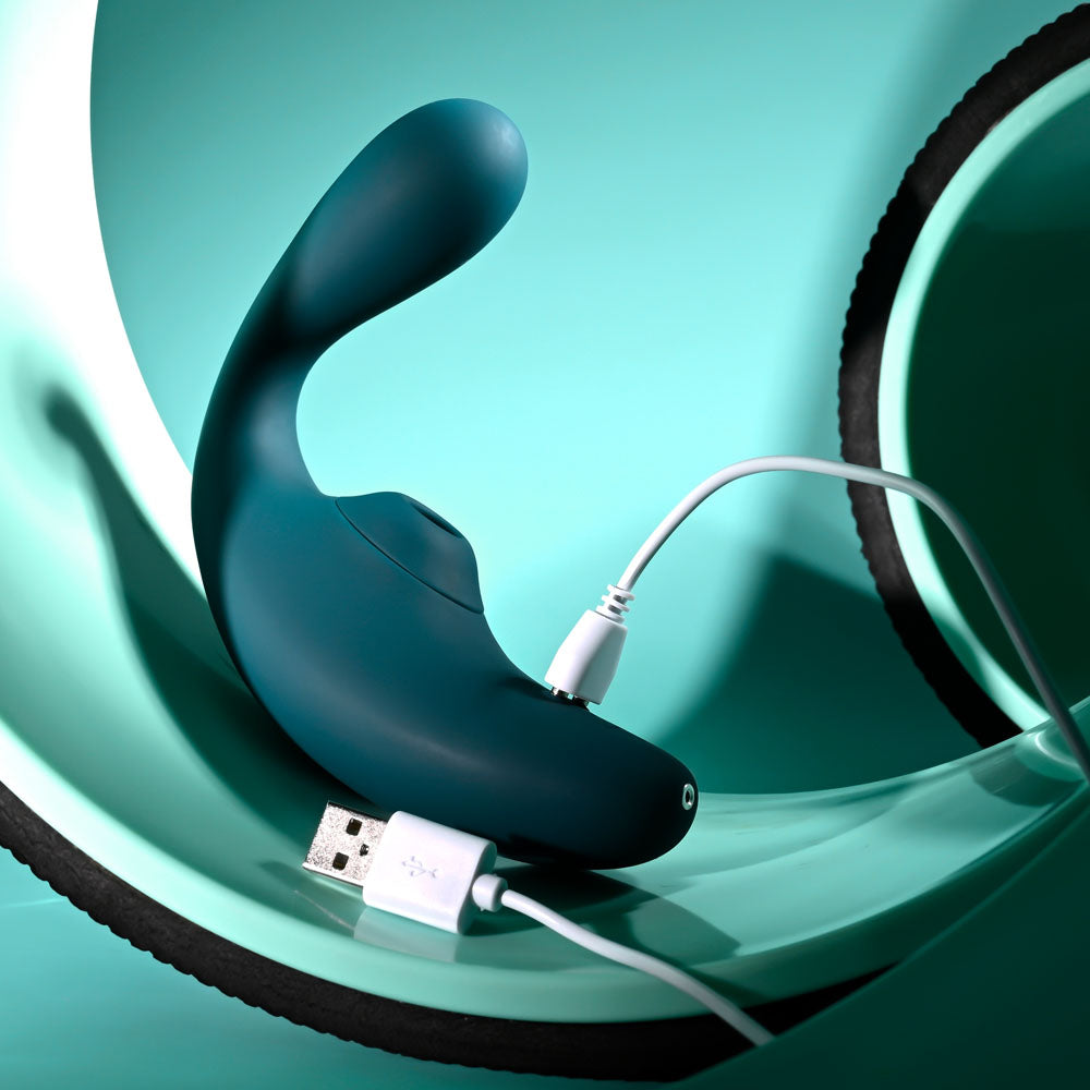 Playboy Pleasure CHARMER -  USB Rechargeable Vibrator with Clitoral Suction charging next to a smooth turquoise surface.