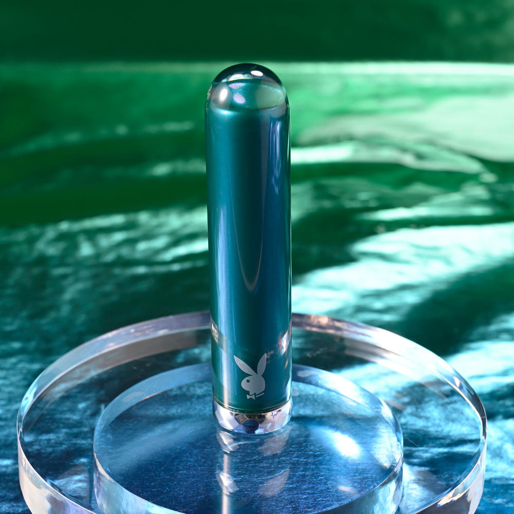 Playboy Pleasure EMERALD -  9.1 cm USB Rechargeable Glass Tipped Bullet on a clear pedestal against a vibrant green background.